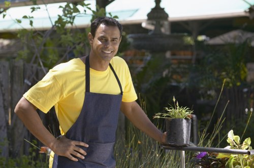 A man with medium skin tone, short dark hair, and a broad smile, dressed in a yellow t-shirt and dark apron, stands outdoors in a garden setting. He is holding a small black pot containing a young plant with green foliage, leaning slightly forward with his right hand on his waist. Behind him, there are various garden elements including plants, shrubs, and trees, with a wooden fence and a garden structure visible in the background. The garden appears well-maintained with lush greenery, and the scene is illuminated by natural daylight, suggesting a clear and sunny day. This image exemplifies gardening activities, such as planting or potting, which are typical services provided by companies like Lawn Mowing Chiswick, that serve gardens in the W4 postcode area, including Chiswick. The overall environment reflects a vibrant, healthy outdoor space suitable for gardening and landscaping maintenance, emphasizing professional outdoor care within a residential garden context.