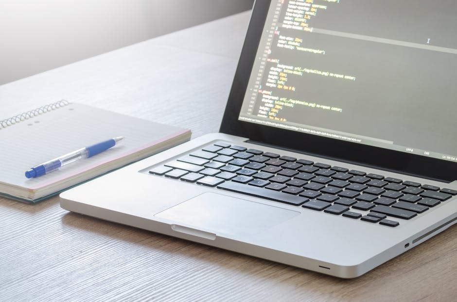 A close-up view of a modern, silver laptop with a black keyboard displaying lines of colorful code on its screen, placed on a light wooden desk. Next to the laptop, there is an open notepad with a blu
