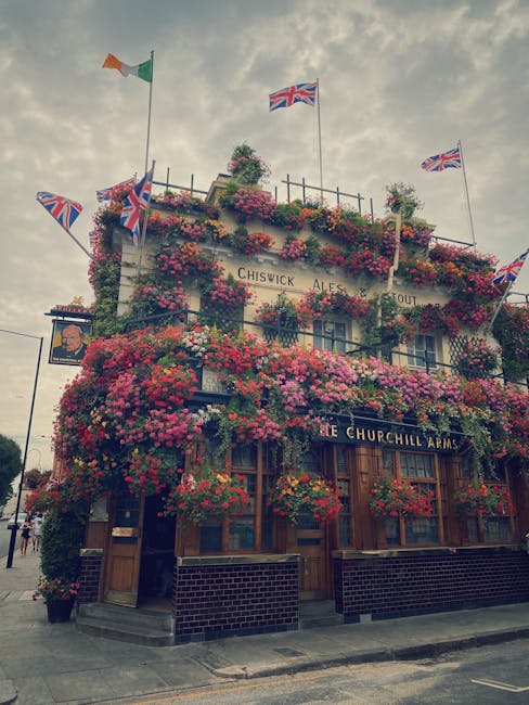 A multi-story pub building, The Churchill Arms, situated on a street corner, extensively decorated with vibrant pink, red, and purple flowers cascading from window boxes and hanging baskets on its fac
