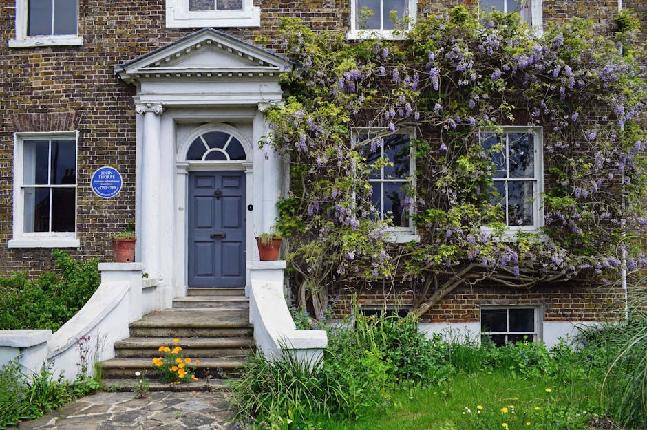 A multi-story pub building, The Churchill Arms, situated on a street corner, extensively decorated with vibrant pink, red, and purple flowers cascading from window boxes and hanging baskets on its fac